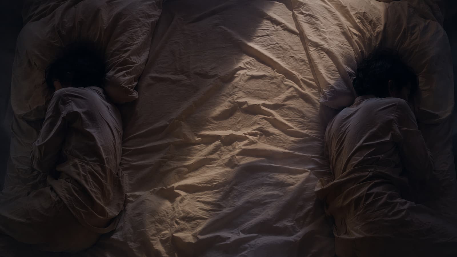 Two figures lying on opposite edges of a bed at dawn, backs turned to each other, warm cream sheets and dim light between them