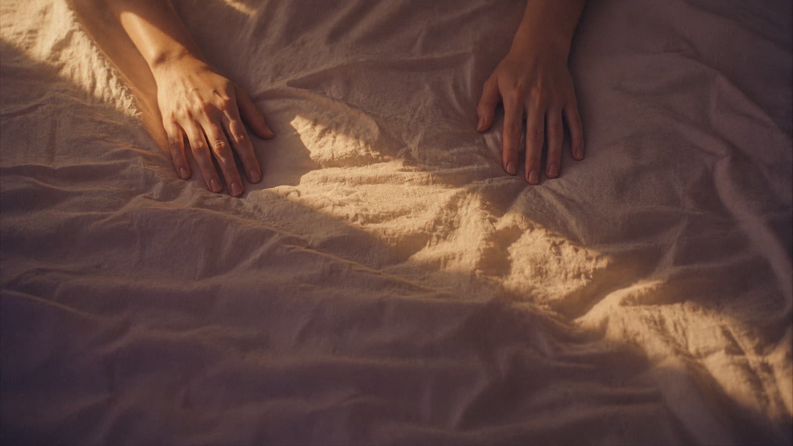 Two hands resting apart on warm-lit unmade bedsheets