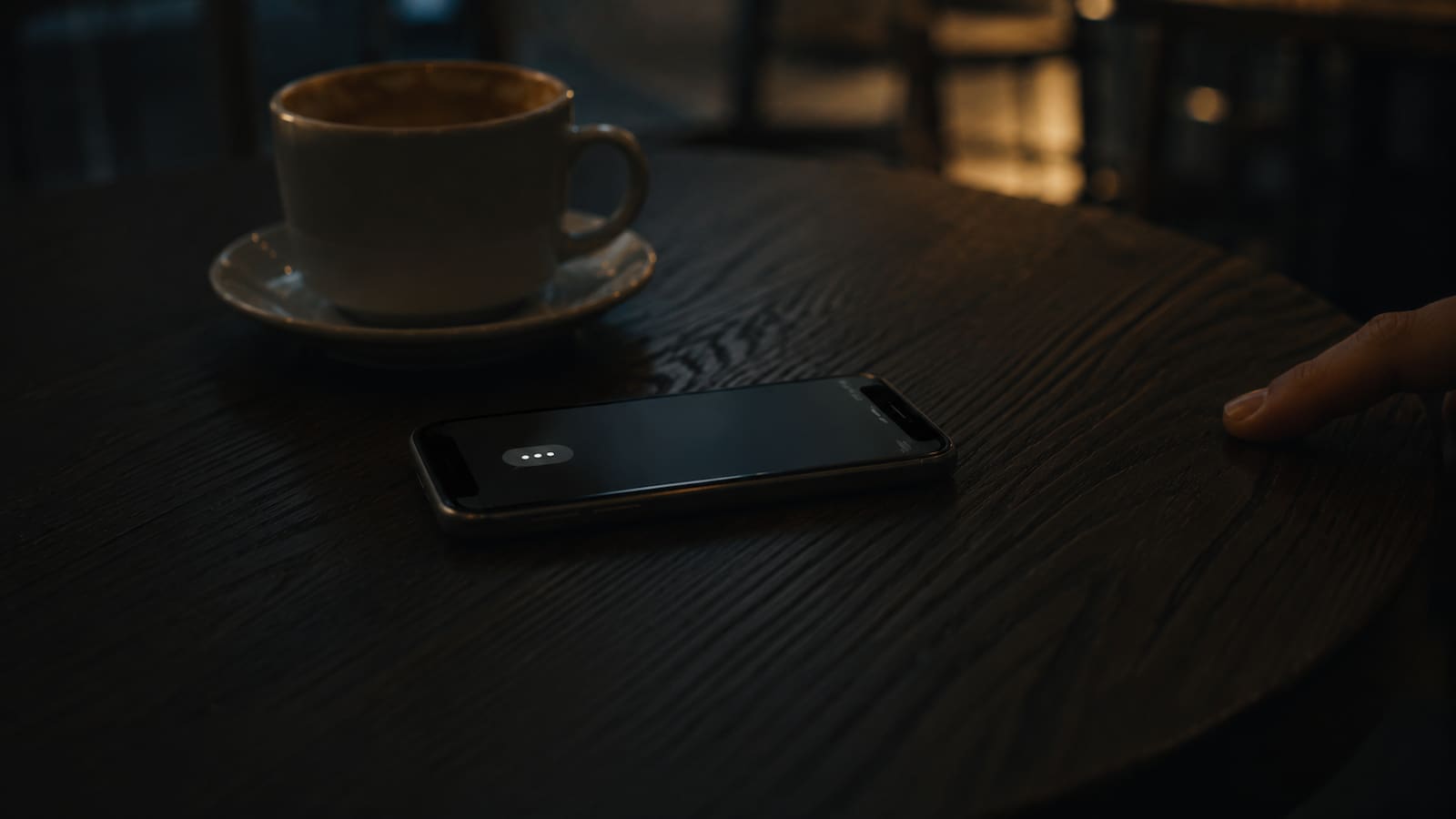 A smartphone face-up on a dark café table showing a typing indicator that has gone still, a half-empty coffee cup behind it, a fingertip resting on the edge of the table