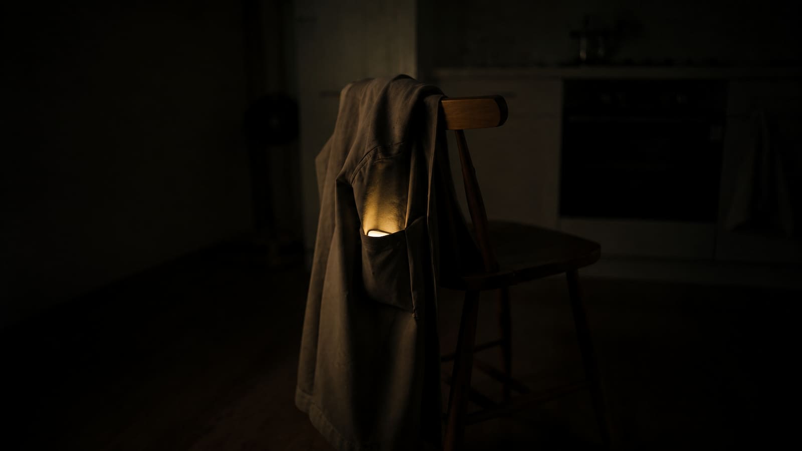 A coat draped on the back of a wooden kitchen chair in a dim apartment at night, a phone glowing softly from inside the pocket