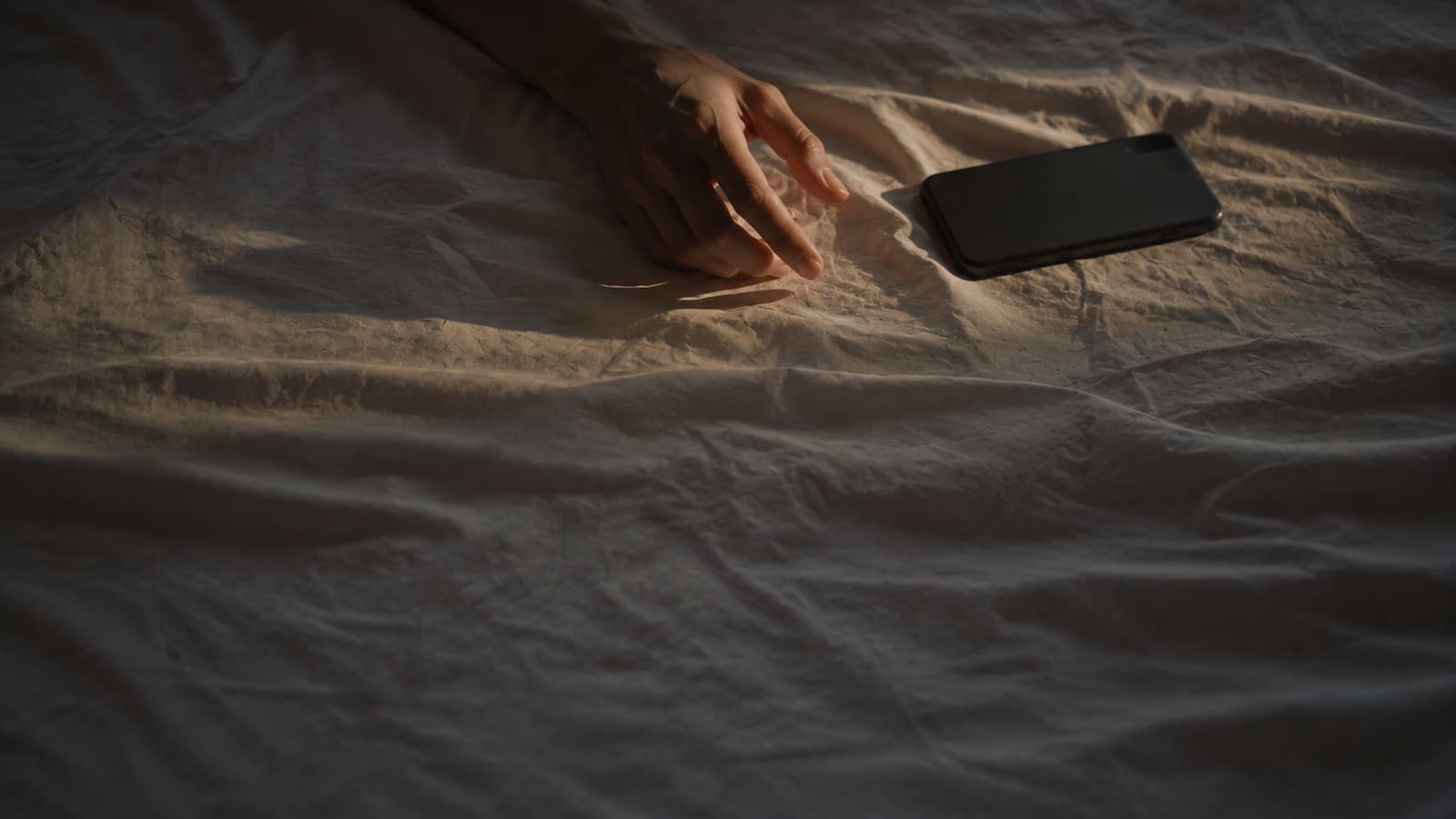 A hand paused mid-reach above a smartphone face-down on a rumpled sheet, warm afternoon light from one side