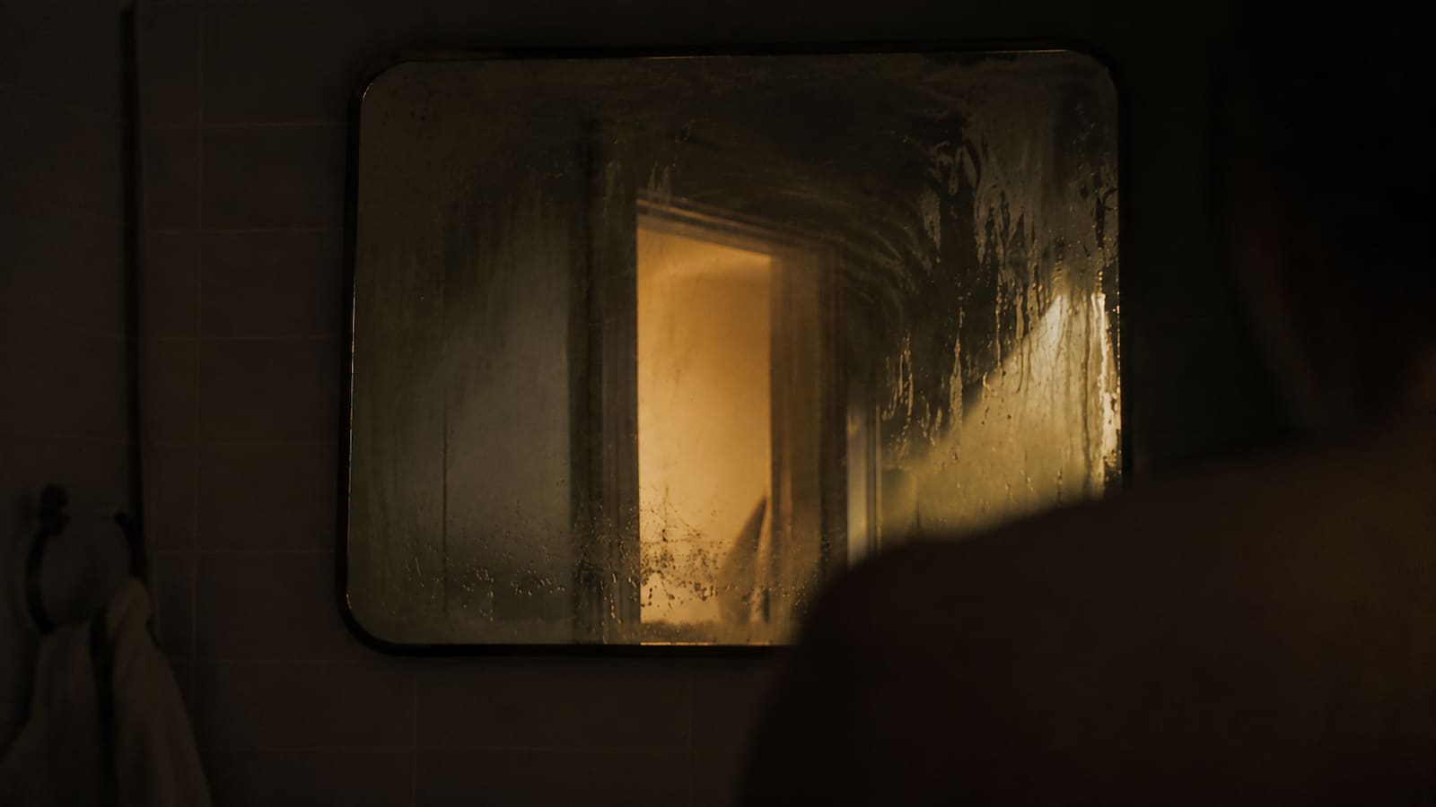 A partially-fogged bathroom mirror at night, warm yellow light spilling from a doorway behind, a blurred shoulder visible at the edge of the reflection
